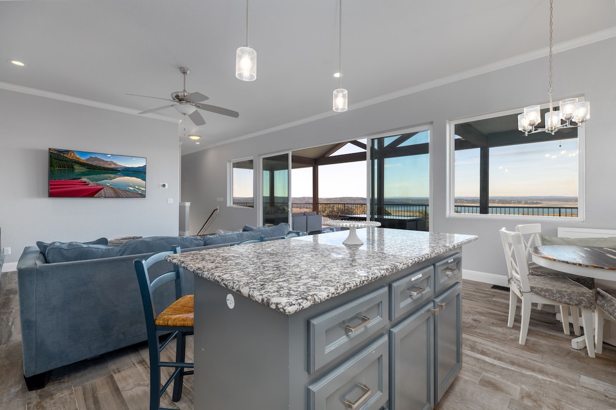 An open-concept living space features a spacious kitchen island topped with light granite and equipped with modern appliances. Large windows offer expansive lake views, allowing natural light to illuminate the area. A cozy seating arrangement is placed nearby, and a dining table is positioned against the wall.