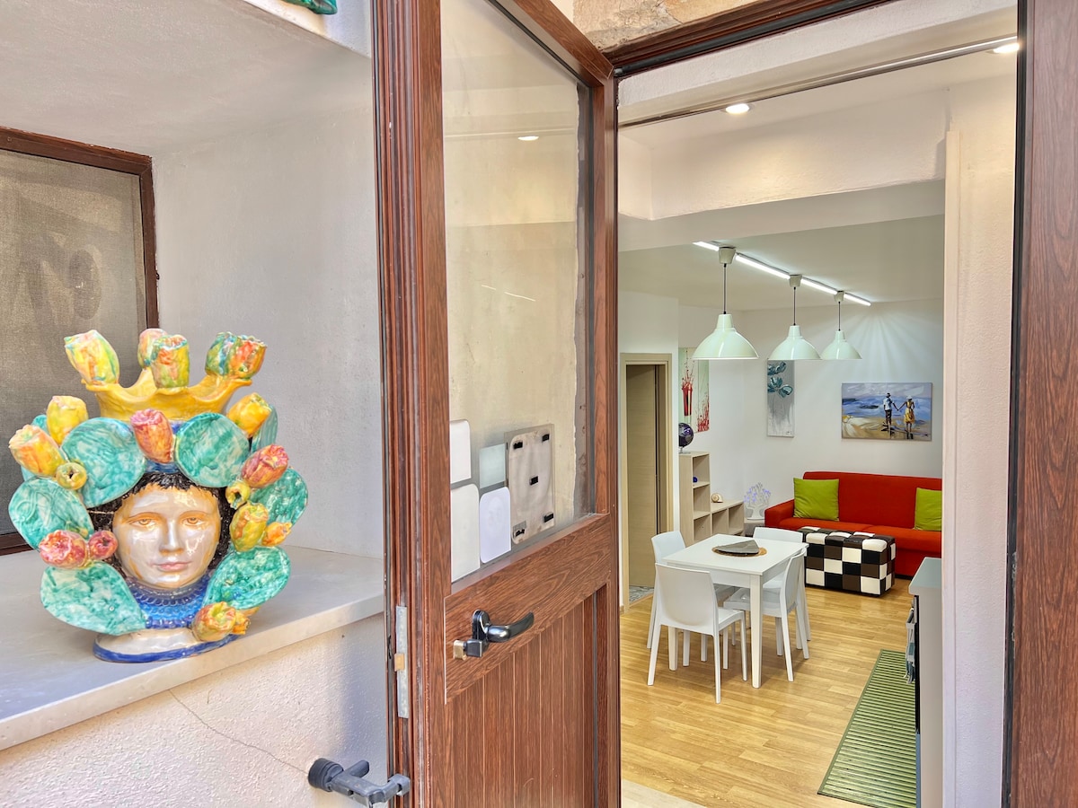 A welcoming entrance is framed by a wooden door, revealing a living area decorated with vibrant artwork and a red sofa. A dining table is set for four, and modern pendant lights illuminate the space, highlighting the varied textures and colors inside.