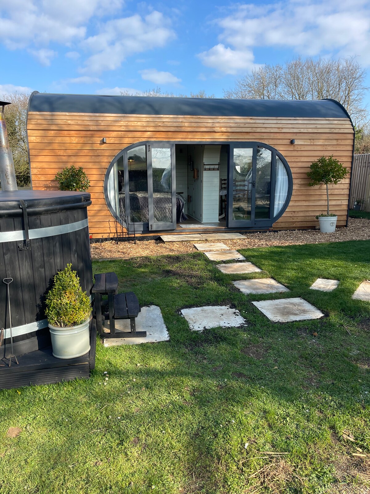 A glamping pod with a curved wooden exterior sits amidst green grass and stone pathways. Large circular glass doors provide an entrance, while two potted plants frame the doorway. A wood-fired hot tub is visible nearby, set against a backdrop of trees and blue sky.