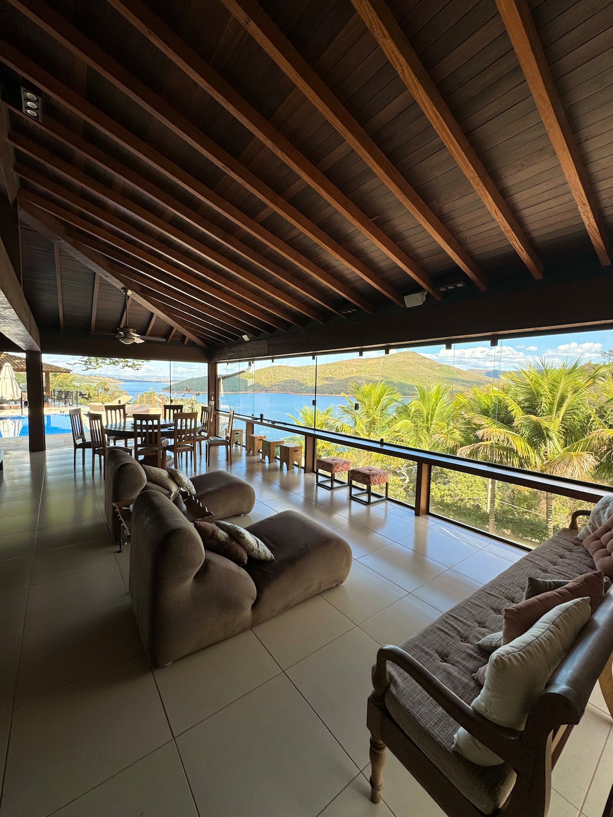 A spacious living area features a wooden ceiling and large glass windows, offering a serene view of the outdoors. Comfortable seating arrangements are present, alongside a dining table. The space is illuminated by natural light, creating a bright and welcoming environment.