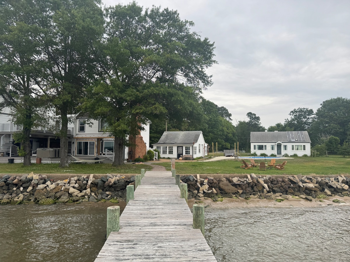 A wooden dock extends from the water towards the cottage, framed by lush greenery. The main house is visible on the left, with the cottage and a fire pit area situated on the right. A peaceful waterfront setting invites relaxation.