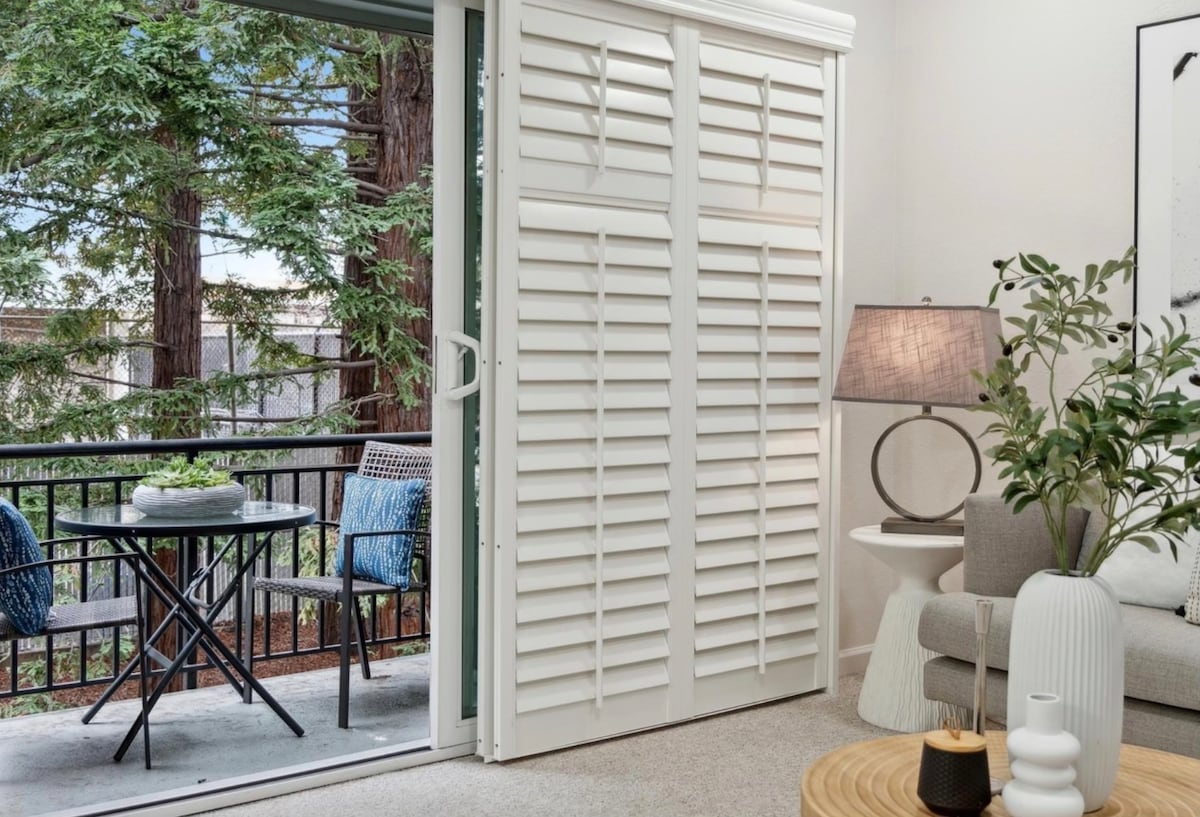 Natural light brightens the living space, where white shutters open onto a private balcony. A small, round black table with two chairs is visible outside, framed by lush green trees, while a round wooden coffee table and a lamp complete the cozy interior.
