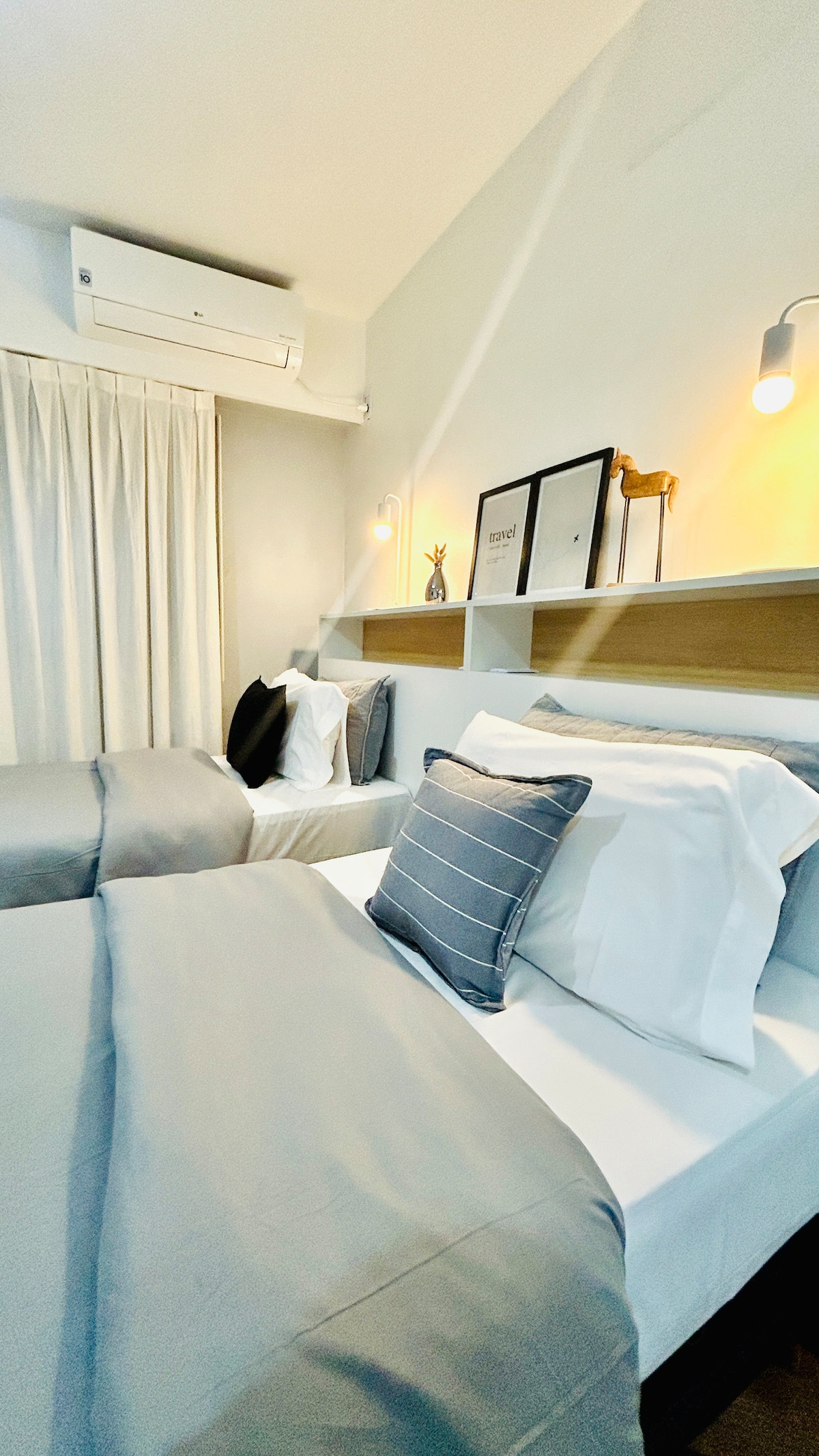 A well-lit sleeping area features two spacious beds with light grey sheets and navy accent pillows. A small shelf above the beds holds decorative items. Natural light enters through a nearby window, shaded by sheer curtains. A split air conditioning unit is mounted on the wall.