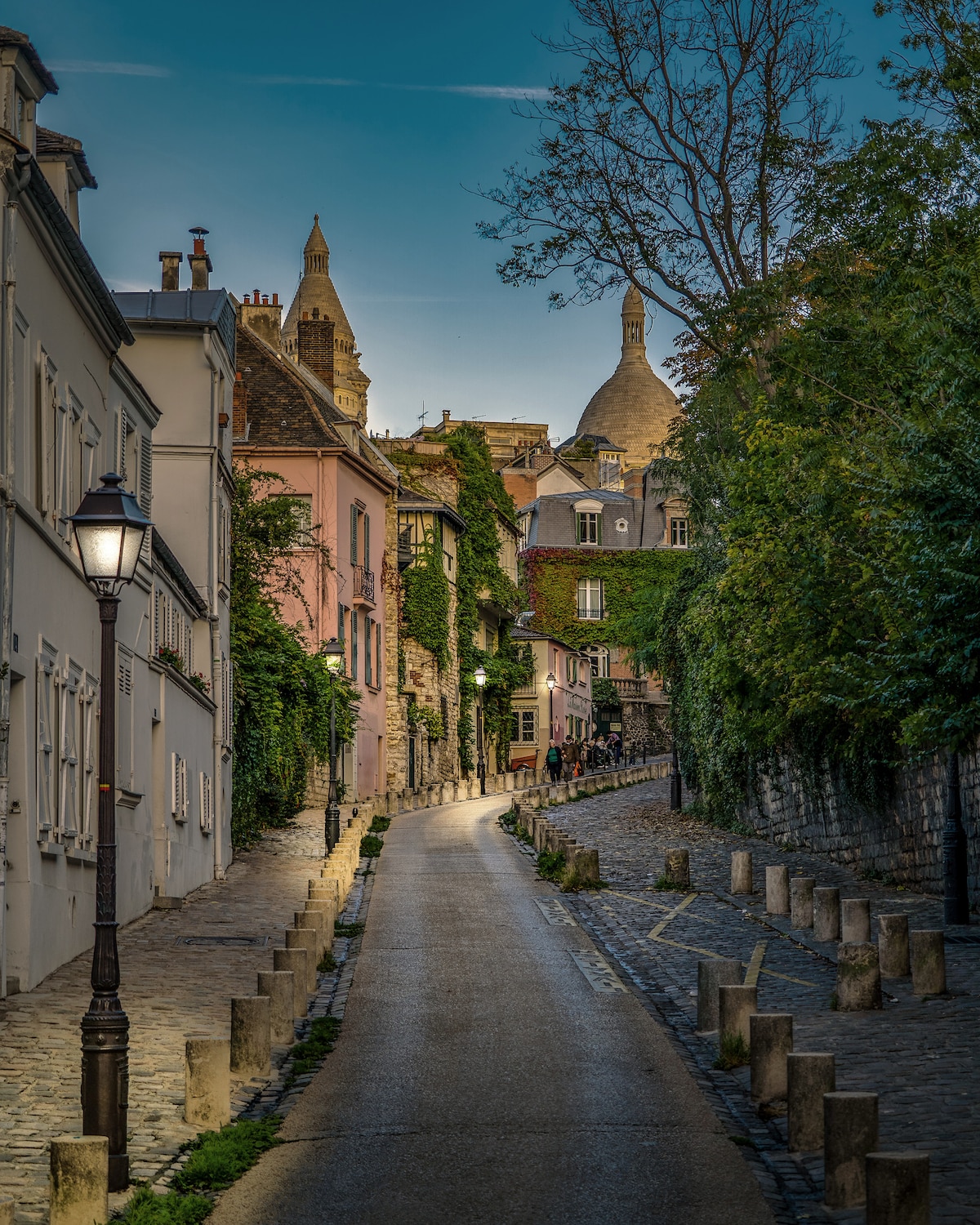 A cobblestone street winds through charming buildings adorned with creeping greenery. Soft street lamps illuminate the pathway, leading towards a hillside where the dome of a church is visible. The scene is set during twilight, creating a serene atmosphere.