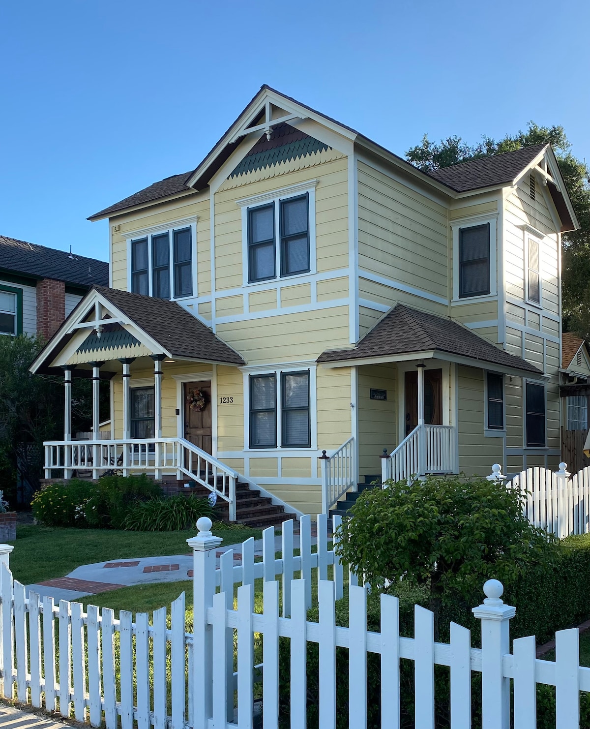 A classic Victorian-style duplex is depicted, featuring a cheerful yellow exterior with white trim. The home has a welcoming porch with decorative elements, complemented by a manicured lawn and a white picket fence. Lush greenery enhances the outdoor space, providing a serene street view.