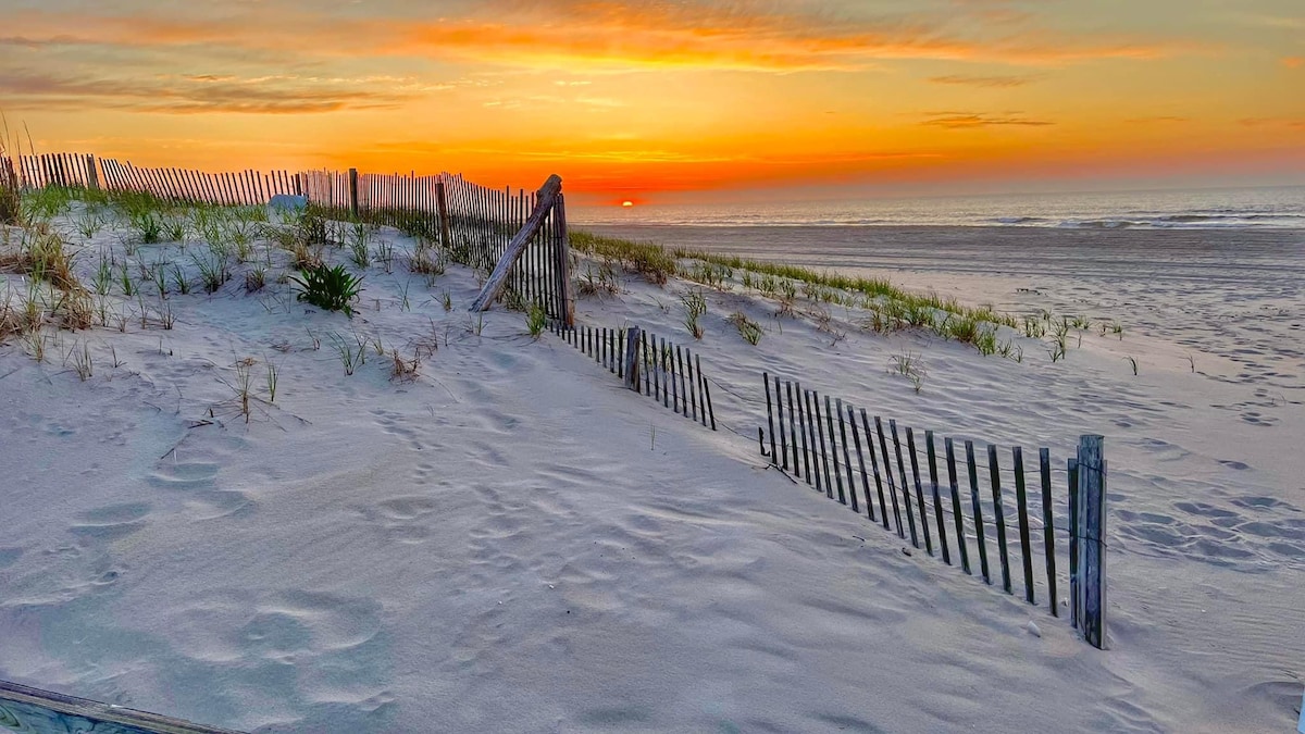 A serene beach scene features soft sand dunes gently sloping towards the ocean, where the sun sets on the horizon. Wooden fencing lines the beach, and tufts of grass peek through the sand, creating a natural and peaceful atmosphere.