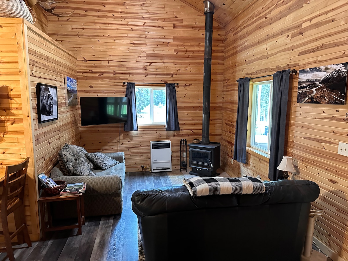A cozy living area features wood-paneled walls and a large window allowing natural light. A plush sofa and a comfortable armchair face a wall-mounted TV, while a small table holds magazines. A stove stands in the corner, enhancing the cabin's warmth.