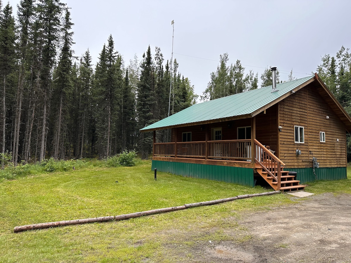 A wooden cabin with a green metal roof is set against a backdrop of dense trees. A wraparound porch features wooden railing and steps leading to the ground. The expansive lawn in front is bordered by natural foliage, offering a sense of seclusion.
