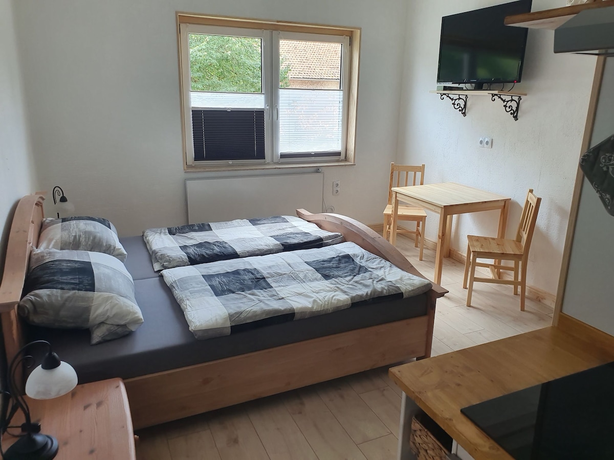 A cozy guest room is presented with a spacious bed fitted with a checkered blanket. A small wooden table and two chairs occupy the corner. An air conditioning unit is mounted in the window, while a television is wall-mounted nearby.