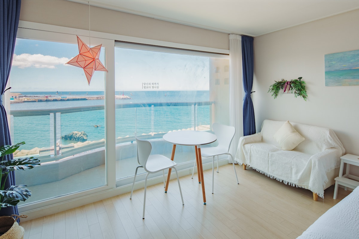 A bright living space features a round dining table paired with two white chairs, positioned near a large window offering a view of the ocean. A light-colored sofa is draped with a soft blanket. Natural light filters in through the sheer curtains.