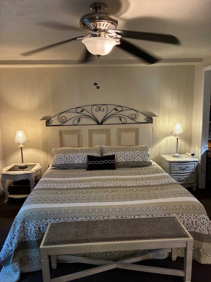 La cama tamaño king en el dormitorio principal con mesas auxiliares y un banco al final de la cama. La cabecera es la puerta principal original reutilizada. El trabajo en hierro es un proyecto de soldadura de Julianne.