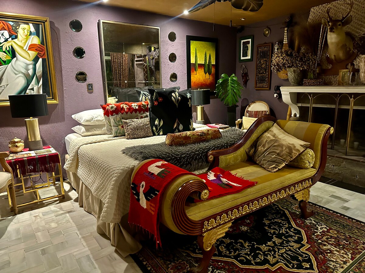 A stylish bedroom is adorned with a mixture of ethnic decor. The spacious bed is layered with various textiles, while a decorative bench provides additional seating. Artwork and unique accents surround the space, complemented by soft lighting from bedside lamps.