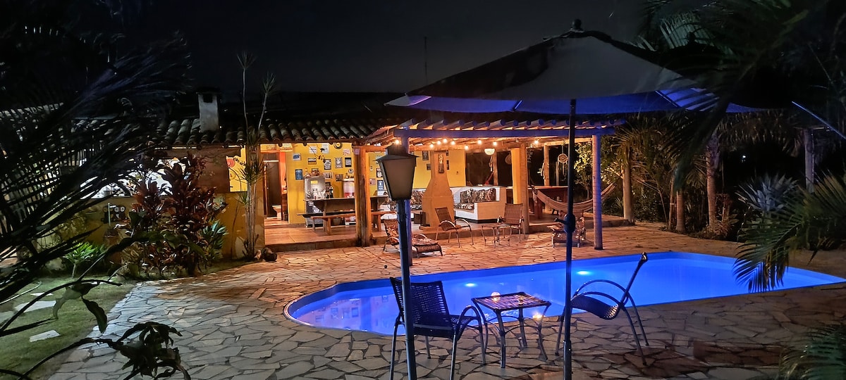 The image captures a nighttime view of the outdoor area, featuring a large illuminated pool surrounded by a stone patio. The house is visible in the background, displaying a warm glow from interior lights. Umbrellas provide shade around comfortable seating areas.