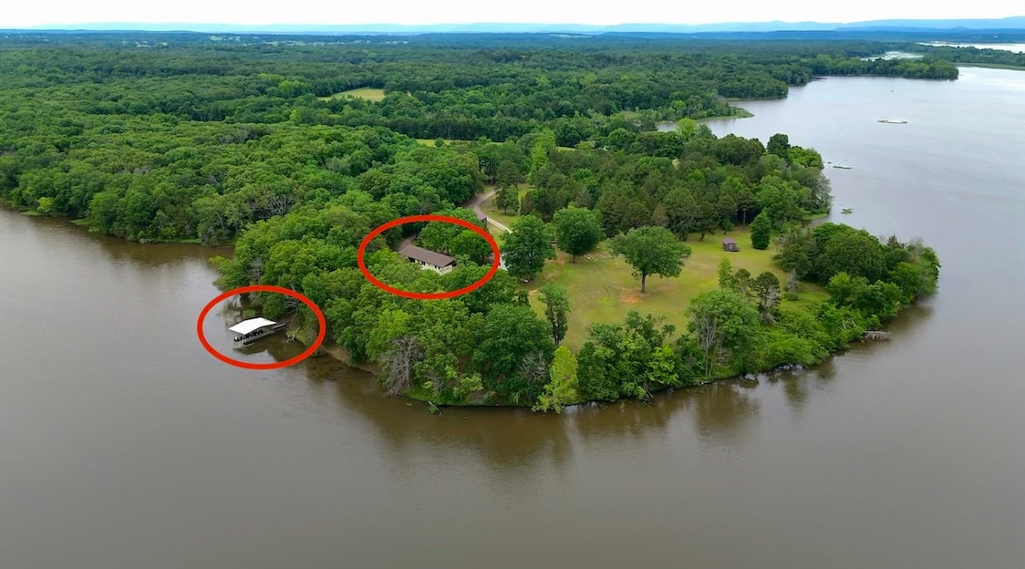 An aerial view captures a serene landscape featuring a spacious property bordered by water. A covered boat dock is visible along the shoreline, alongside a house set amongst greenery. Surrounding trees enhance the natural setting, providing a sense of privacy and tranquility.