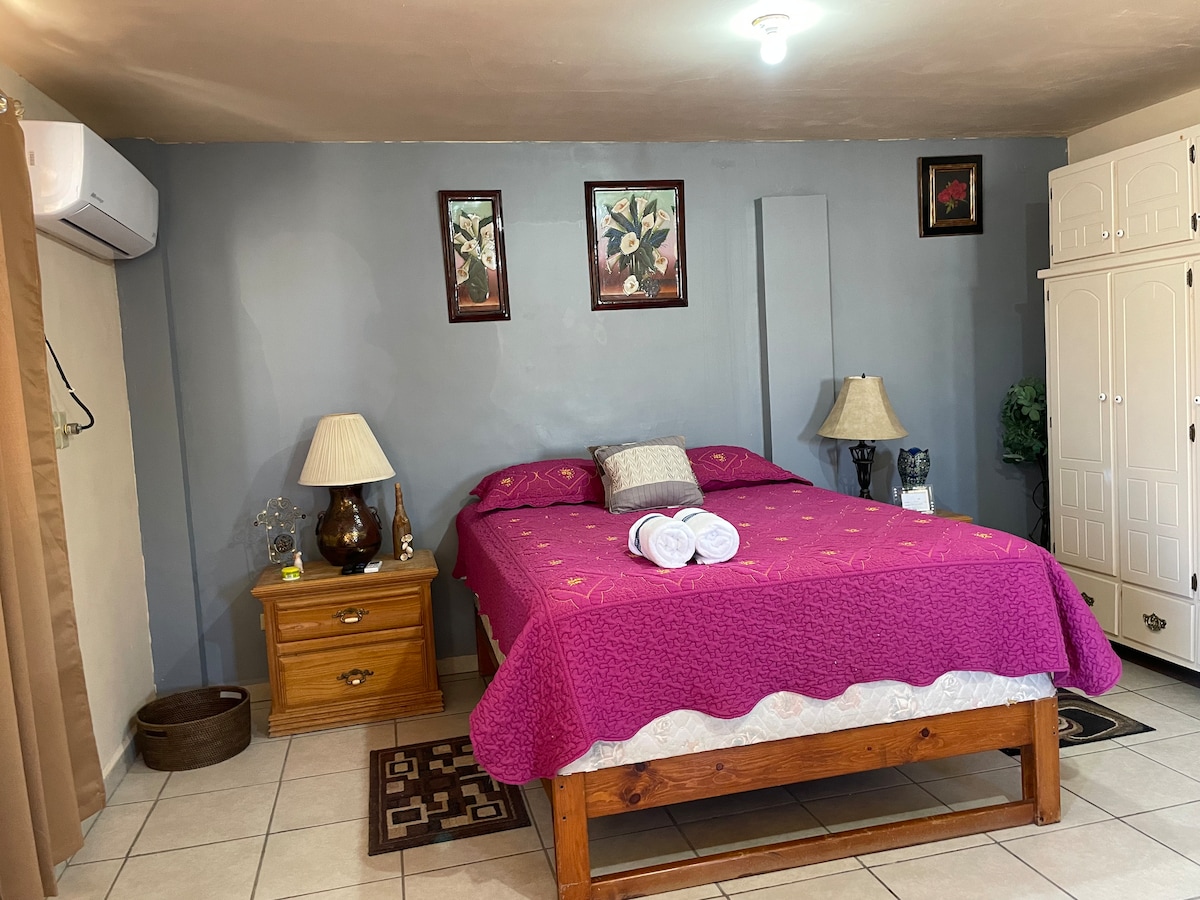 A spacious sleeping area is highlighted by a large bed dressed in a pink quilt. Nightstands with lamps are positioned on either side. A decorative wall art piece and a closet with multiple compartments can be seen. Soft lighting adds to the comforting ambiance.