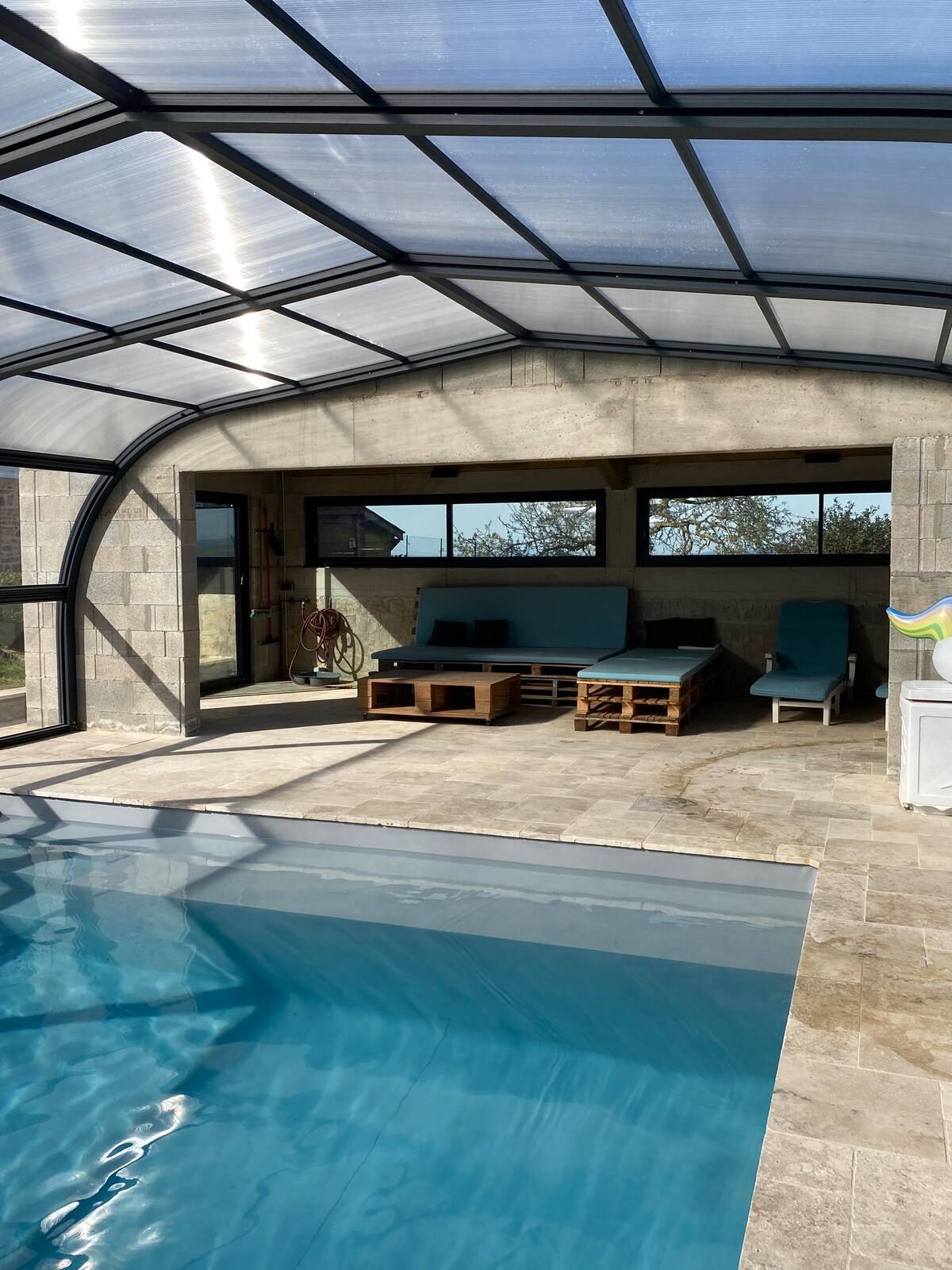 An indoor pool area is enclosed by a transparent roof, allowing natural light to brighten the space. Loungers are arranged nearby on a tiled surface, with a pool visible in the foreground. Large glass doors lead to an outdoor area, enhancing the connection to nature.