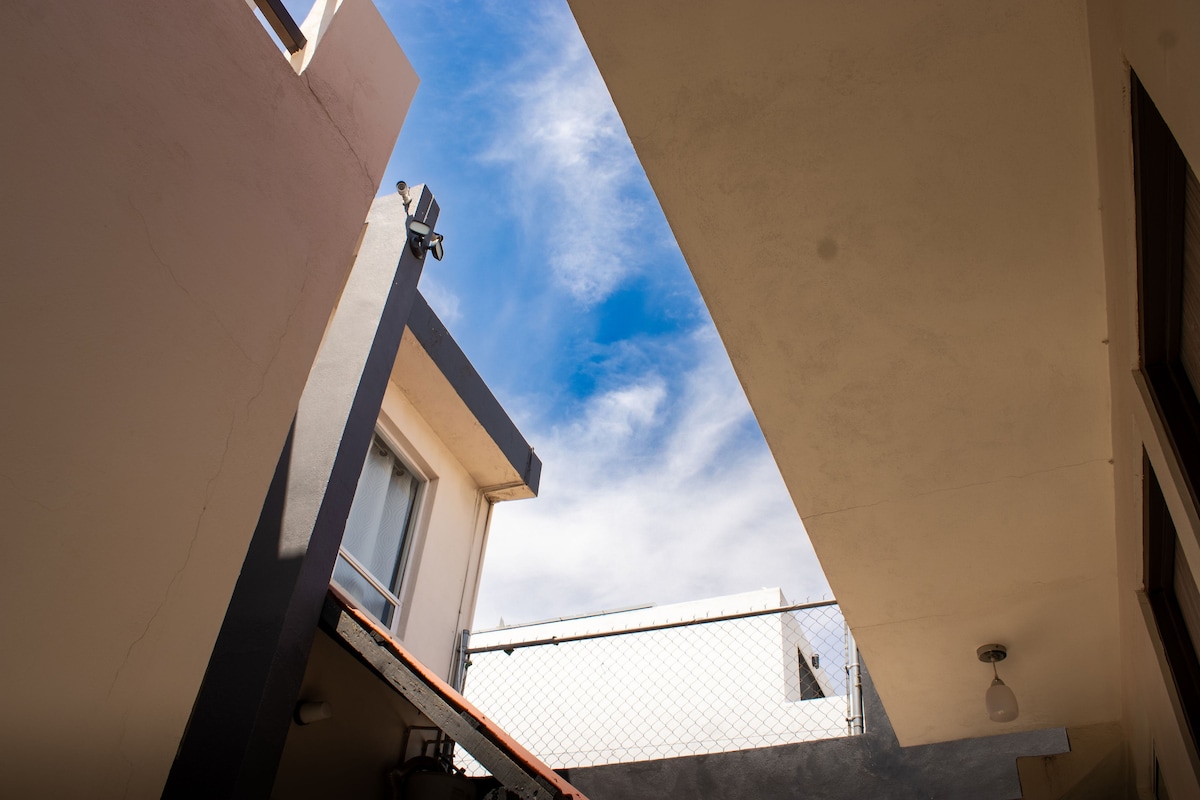 A view of the sky is presented through the spaces between two buildings, showcasing a mix of blue and white clouds. Natural light filters in, illuminating the nearby surfaces. The image captures the architectural outlines and textures of the surrounding structures.