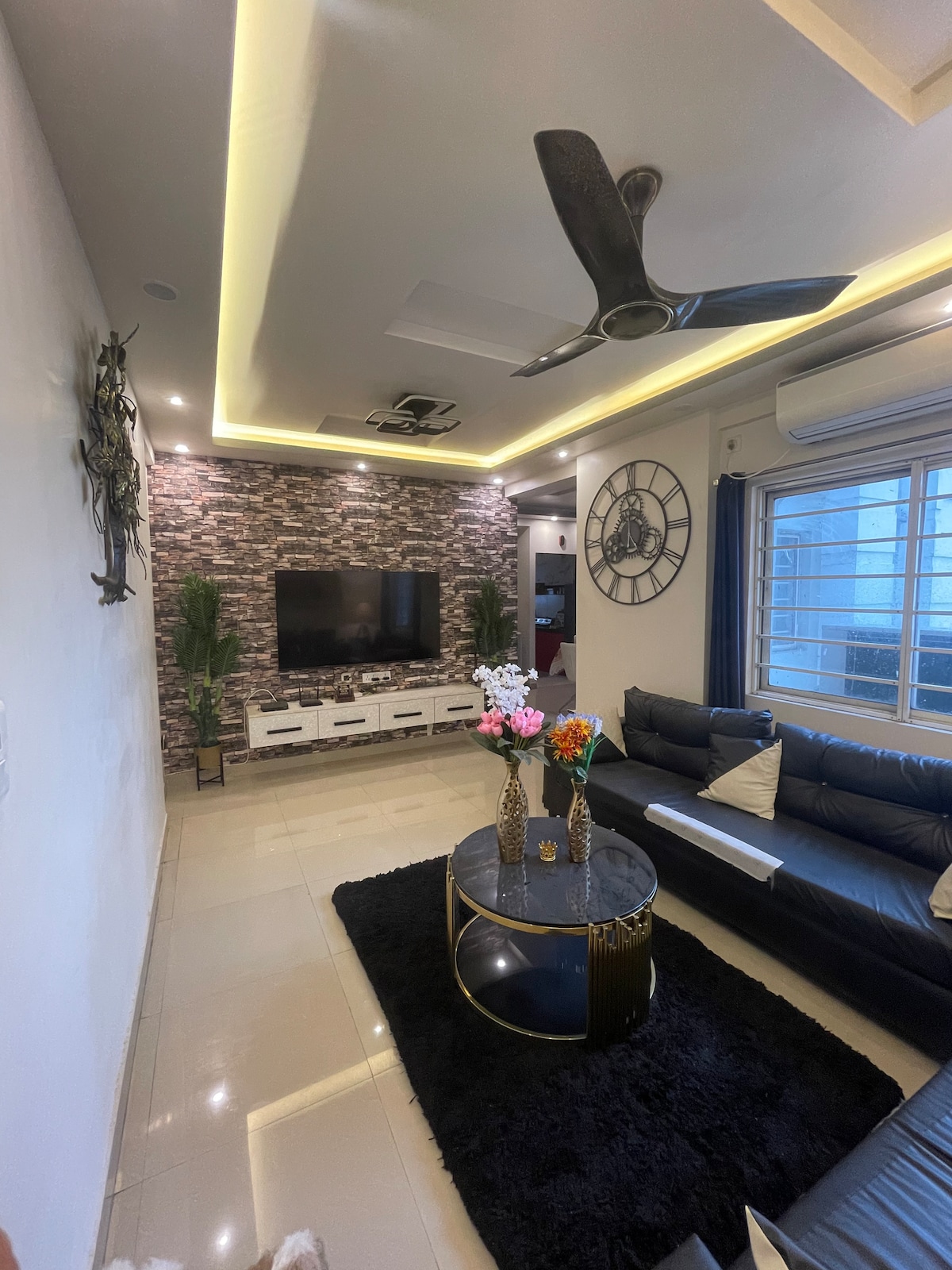 The living room features a modern design highlighted by a dark accent wall and stylish furnishings. A large television is mounted on the wall, complemented by a circular coffee table with decorative vases. The area is illuminated by recessed lighting and a ceiling fan.