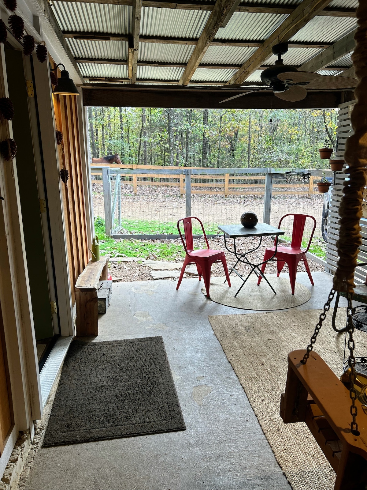 An outdoor seating area is seen, featuring a small round table with two red chairs. The space offers views of a fenced grassy yard, surrounded by trees, with natural light filtering in through the open entryway.
