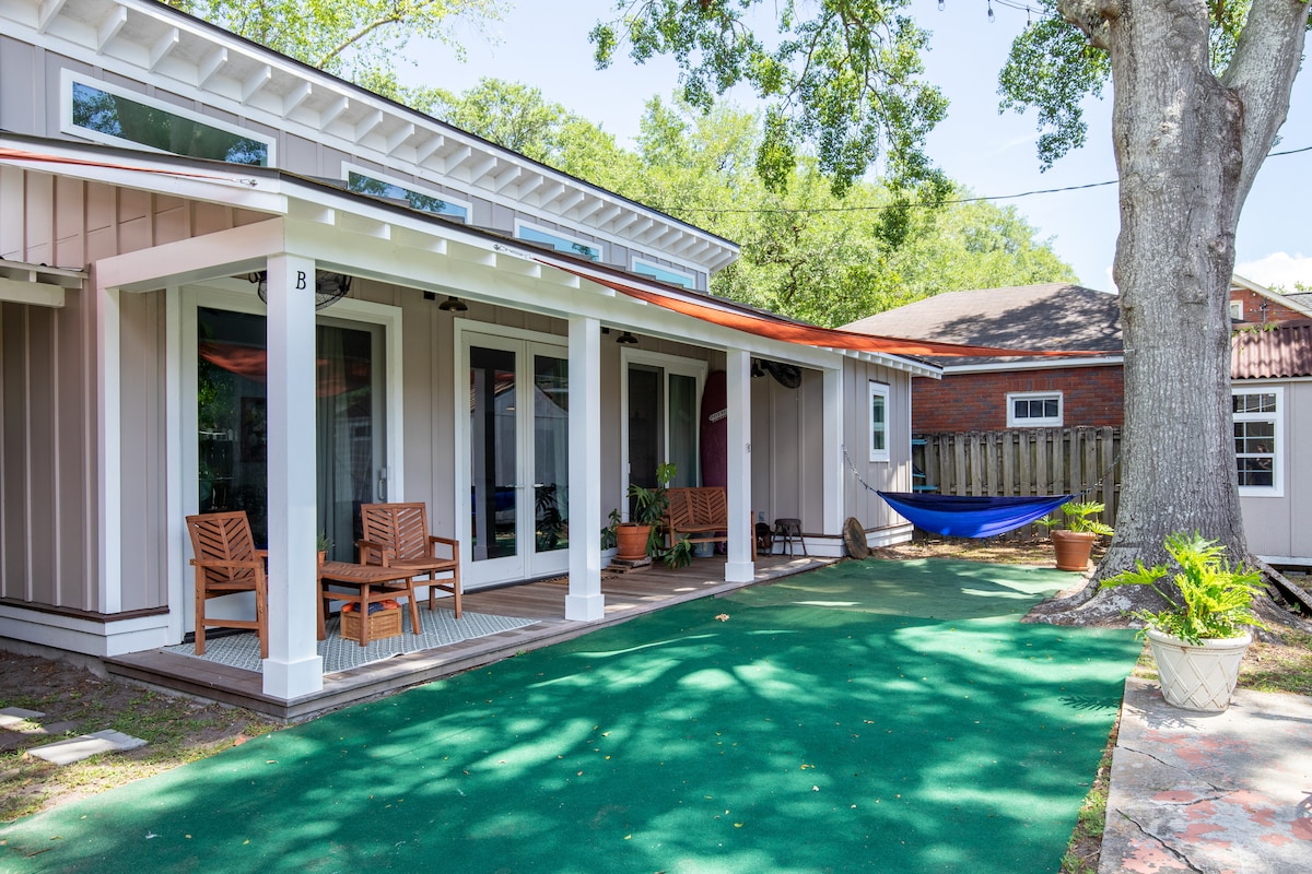 An outdoor seating area features handcrafted wooden chairs placed on a green surface, with sunlight filtering through the surrounding trees. A hammock is seen tucked between two supports, adding a relaxing touch to the open space. Large glass doors provide views into the studio's interior.