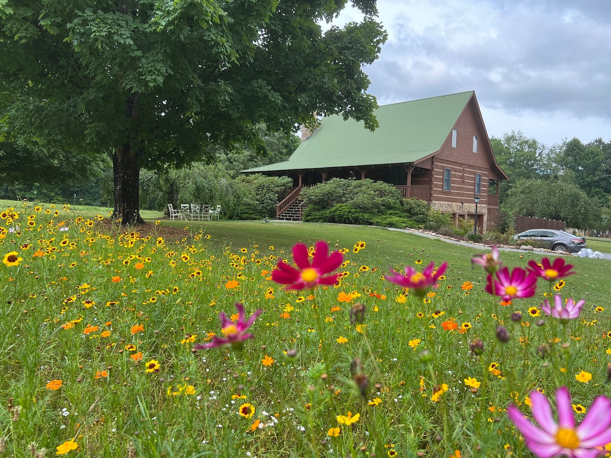 A log home with a green metal roof is surrounded by a vibrant flower garden featuring assorted wildflowers in shades of pink, yellow, and orange. A spacious porch is visible, inviting guests to enjoy the natural landscape and peaceful surroundings.