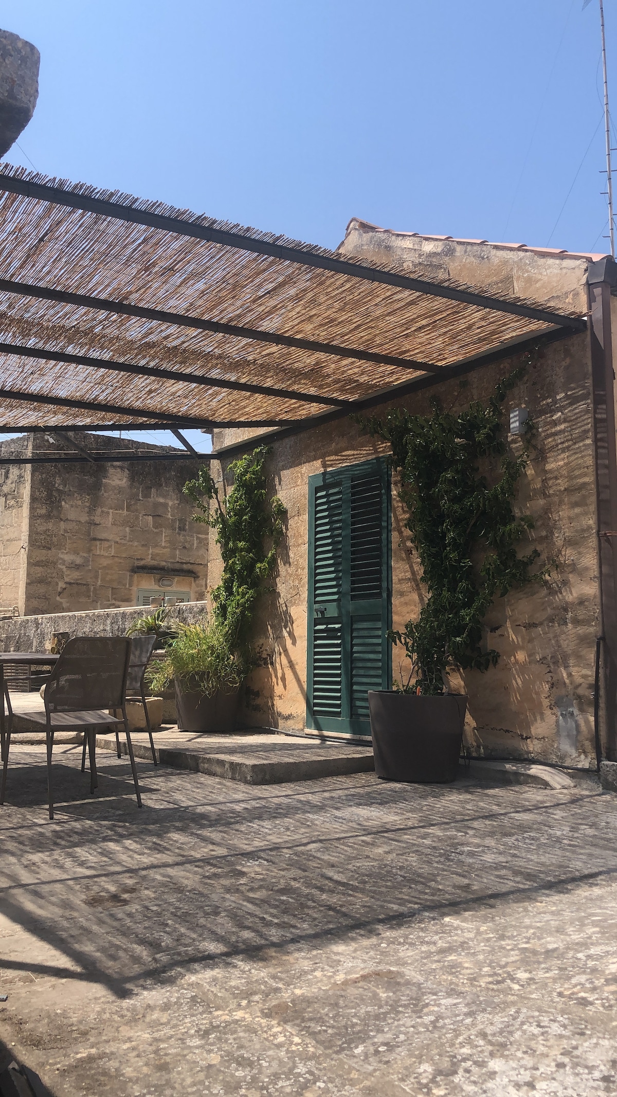 An outdoor terrace is showcased, featuring a natural fiber canopy providing shade. Green plants are placed in large pots, and a few metal chairs are arranged on the stone floor. A green shuttered door adds a rustic charm, complementing the sunlit surroundings.