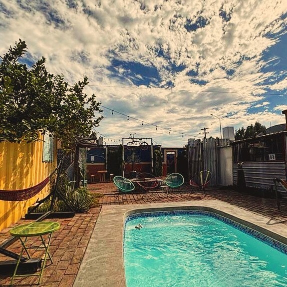 A tranquil pool area features a clear blue pool surrounded by a brick patio. Colorful chairs and a hammock are positioned nearby, set against a backdrop of a lively sky filled with clouds. String lights hang above, creating a relaxed atmosphere.