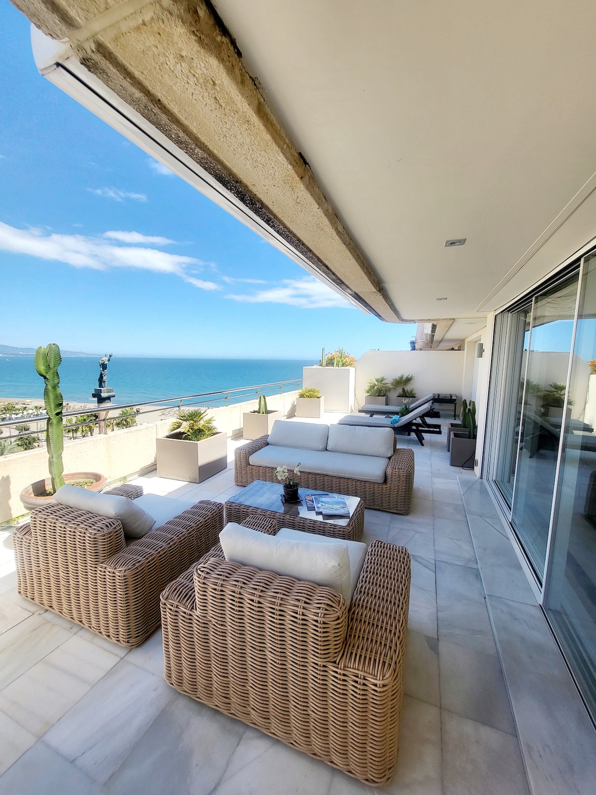 An outdoor terrace is presented, featuring comfortable wicker seating arranged around a small table. A clear view of the ocean is visible, with a bright blue sky and distant mountains in the background. Potted plants are placed around, enhancing the space's greenery.