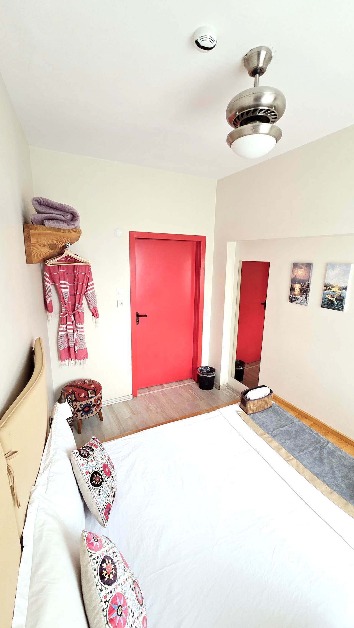 A cozy room features a spacious bed dressed in white linens, complemented by decorative throw pillows. A vibrant red door leads to the hallway, while a stylish ceiling light illuminates the space. A wooden shelf holds towels, and a mirror reflects part of the room.