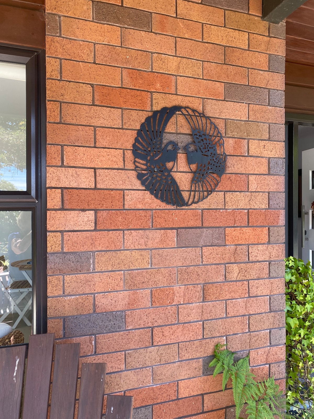 A decorative wall hanging of two birds is affixed to a brick wall, showcasing intricate detailing. The texture of the red and brown bricks contrasts with the dark metal of the artwork. Nearby, greenery adds a natural element, complementing the welcoming entryway.