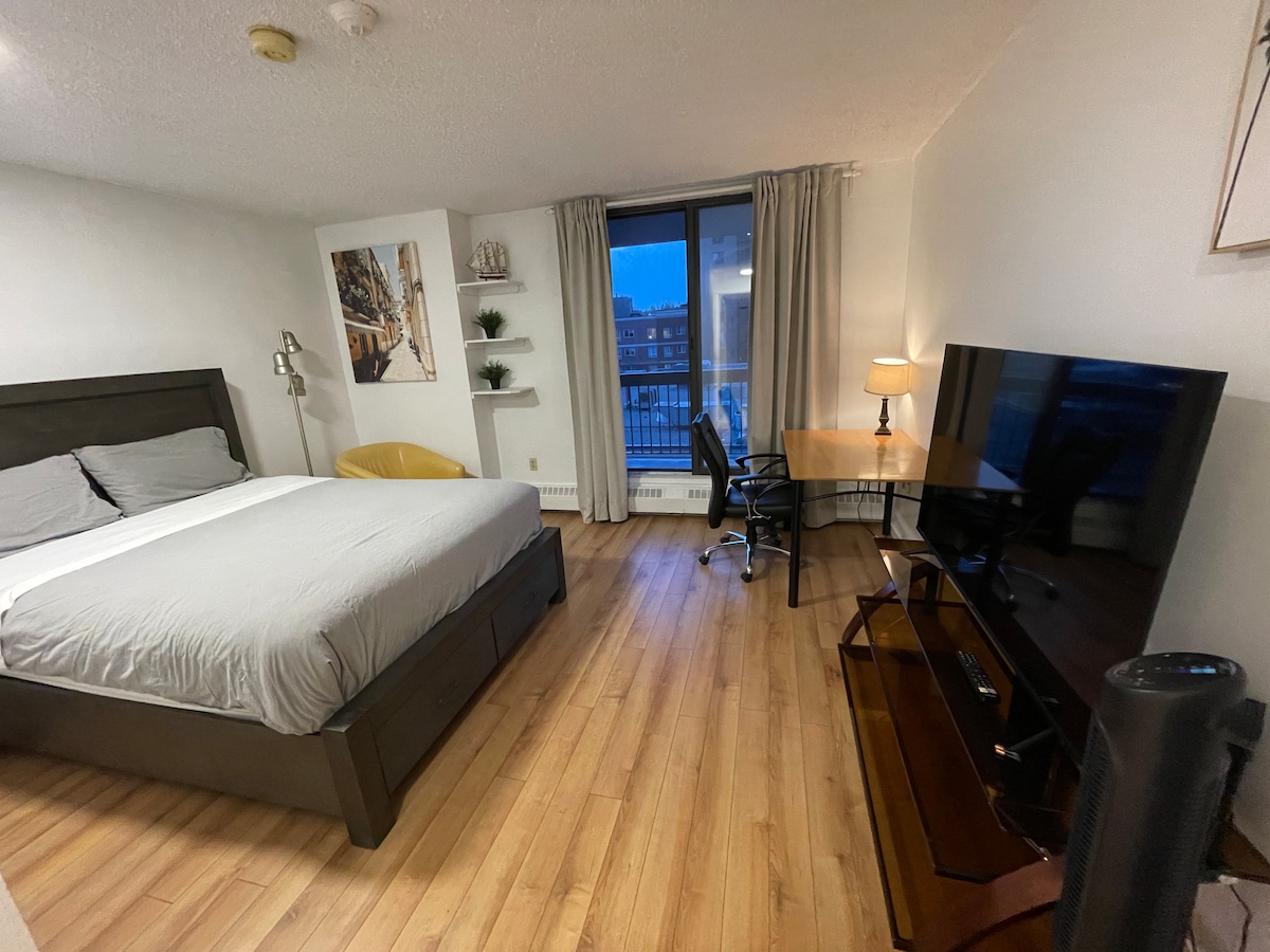 The uncluttered living space features a spacious bed with soft linens, an entertainment area with a TV, and a work desk beside a large window. Natural light enters through the window, creating a bright atmosphere. A chair and lamp provide additional comfort and functionality.