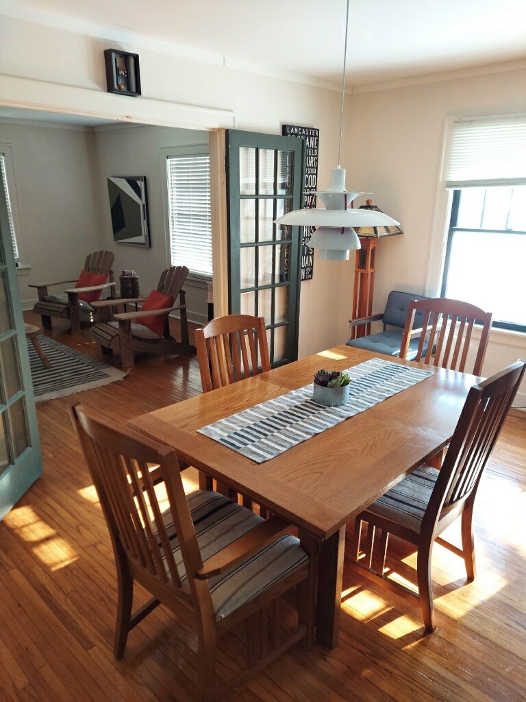 A dining area features a wooden table surrounded by four chairs, with sunlight illuminating the space. A striped runner decorates the table, which is set against a backdrop of bright, airy windows and a comfortable seating area visible through a nearby doorway.