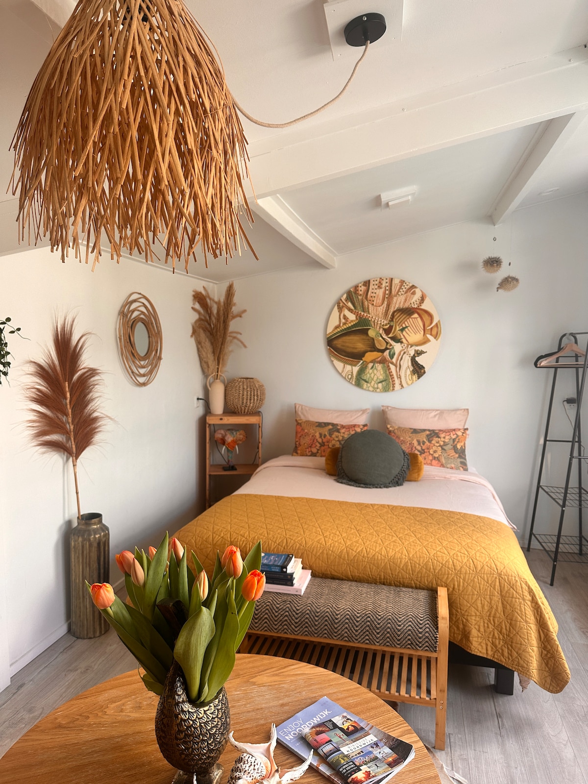 The sleeping area features a spacious bed dressed in warm yellow bedding. A decorative wall hanging and various plant elements contribute to the cozy ambiance. A wooden table holds a vase of tulips, while a stylish light fixture enhances the overall design.
