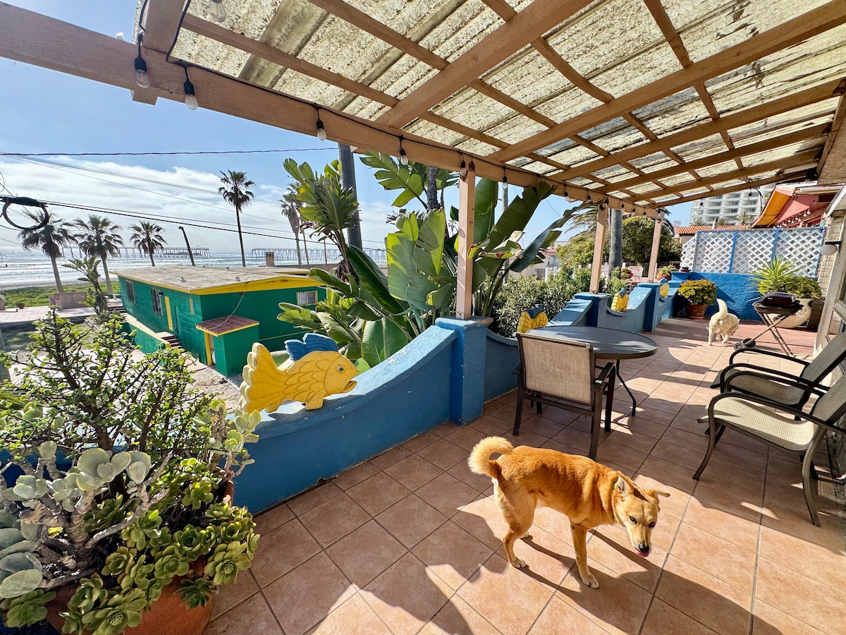 An outdoor patio area is presented, featuring a tiled floor and covered by a wooden pergola. Lush plants and decorated walls emphasize the tropical setting. A dining set is visible, with a friendly dog walking nearby. The ocean view is complemented by palm trees in the background.