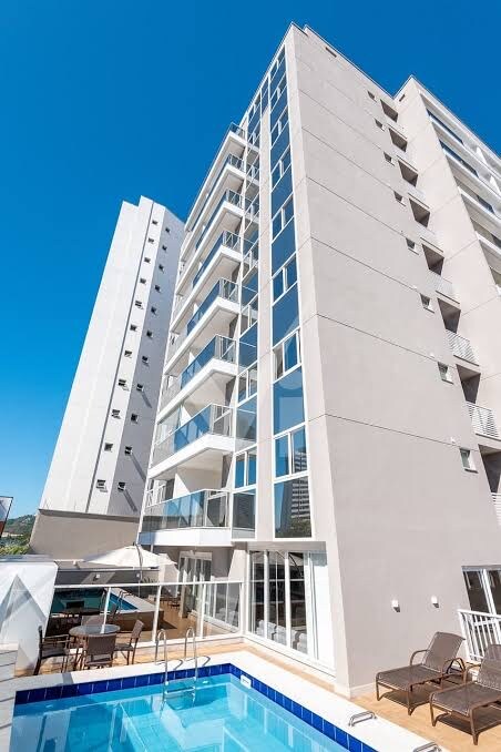 The modern building showcases a sleek façade with large glass windows, allowing for ample natural light. A serene pool is situated at the ground level, accompanied by sun loungers and umbrellas, creating a relaxed outdoor area for residents.
