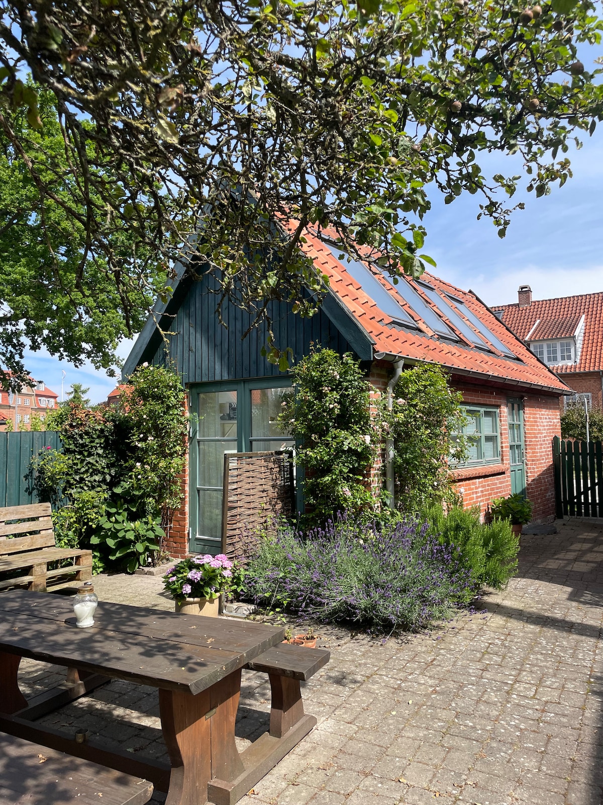 The exterior of a charming red-brick building is showcased, featuring green accents and a sloped roof. Lush greenery decorates the facade, while a wooden table and benches are available in the outdoor space. Flower planters add a touch of color to the area.