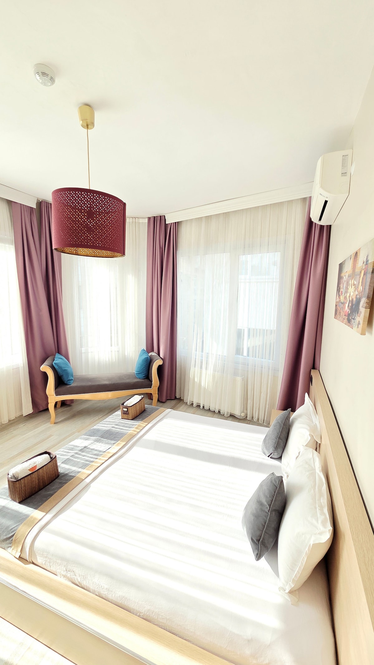 A bright room is adorned with sheer white curtains that allow natural light to fill the space. A spacious bed with soft linens is complemented by decorative pillows. A comfortable wooden seating area is included, along with a simple light fixture above, creating a relaxed atmosphere.