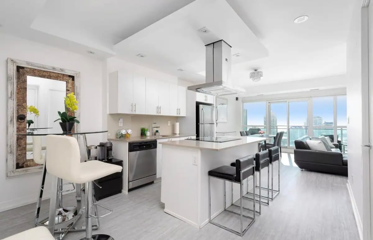 A modern kitchen features sleek white cabinetry and stainless-steel appliances, complemented by stylish bar stools at the island. Large windows provide abundant natural light and offer a view of the city skyline beyond, while a mirror enhances the sense of space.