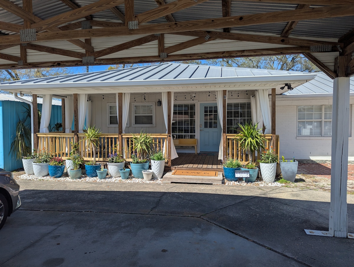 The exterior of the property features a welcoming porch adorned with potted plants. Light-colored curtains drape from the porch, creating a sense of privacy. A blue door invites guests inside, while the surrounding area showcases a paved driveway and carport. Natural light brightens the scene.