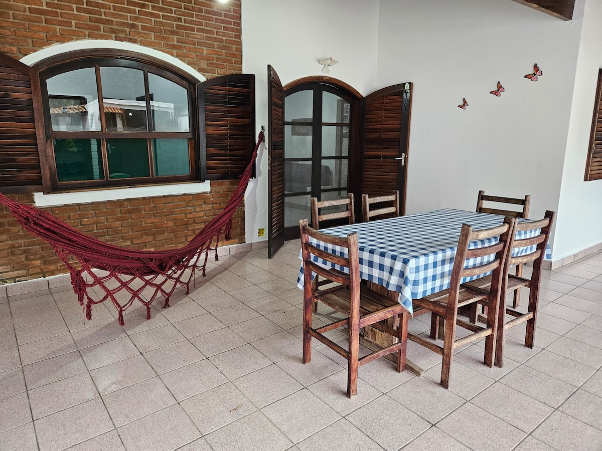 An open dining area is visible, featuring a wooden table with six chairs and a checkered tablecloth. A red hammock is draped between two walls. Natural light enters through two large windows, with wooden frames adding a touch of warmth to the space.