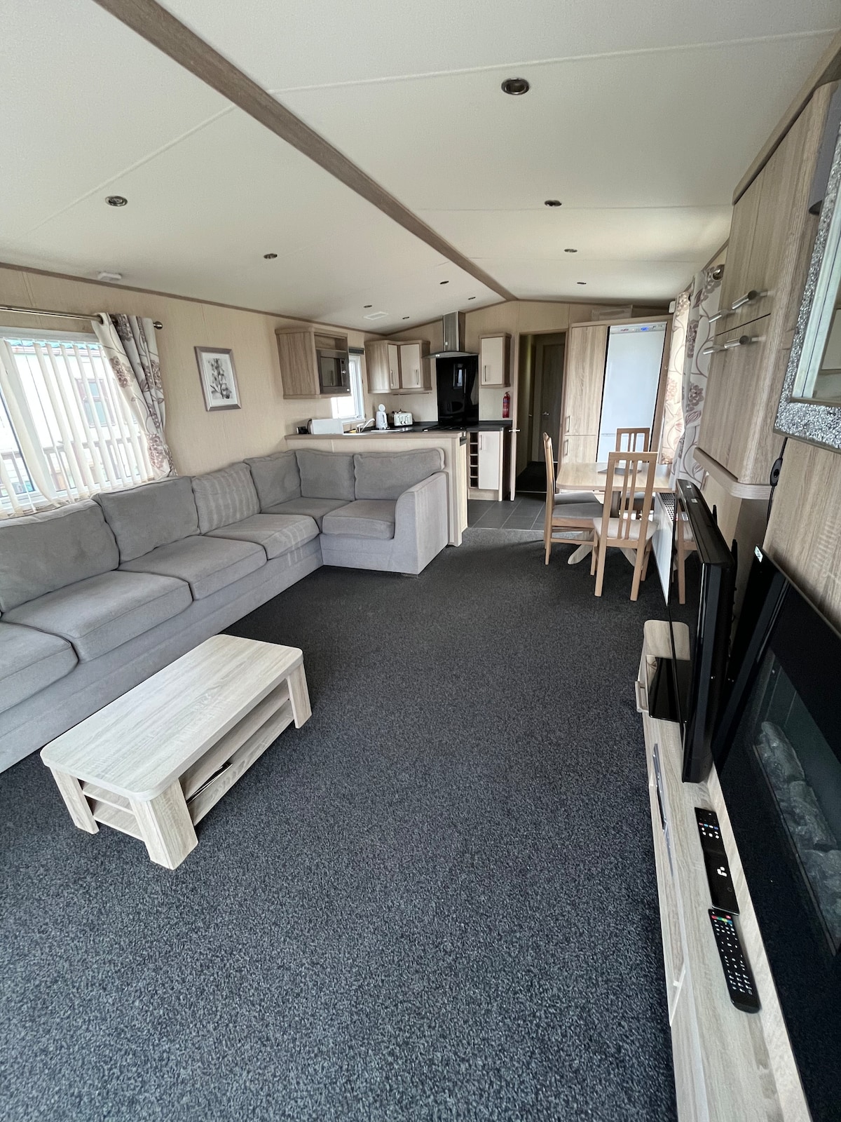 A spacious living area features a light gray sectional sofa paired with a wooden coffee table. The open-plan design is complemented by a kitchen area with modern appliances. Two televisions are positioned on a media console, and natural light enters through large windows covered with patterned curtains.
