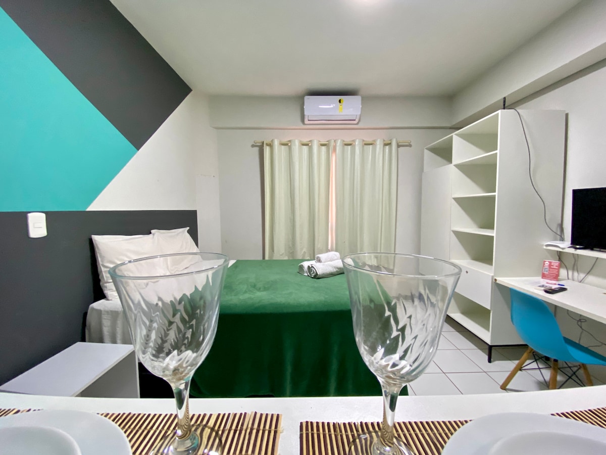 The image features a well-lit room with a spacious bed draped in a green cover, complemented by light curtains. In the foreground, two crystal-clear glasses rest on a dining setup, while a minimalist workspace with a blue chair and television is visible in the background.