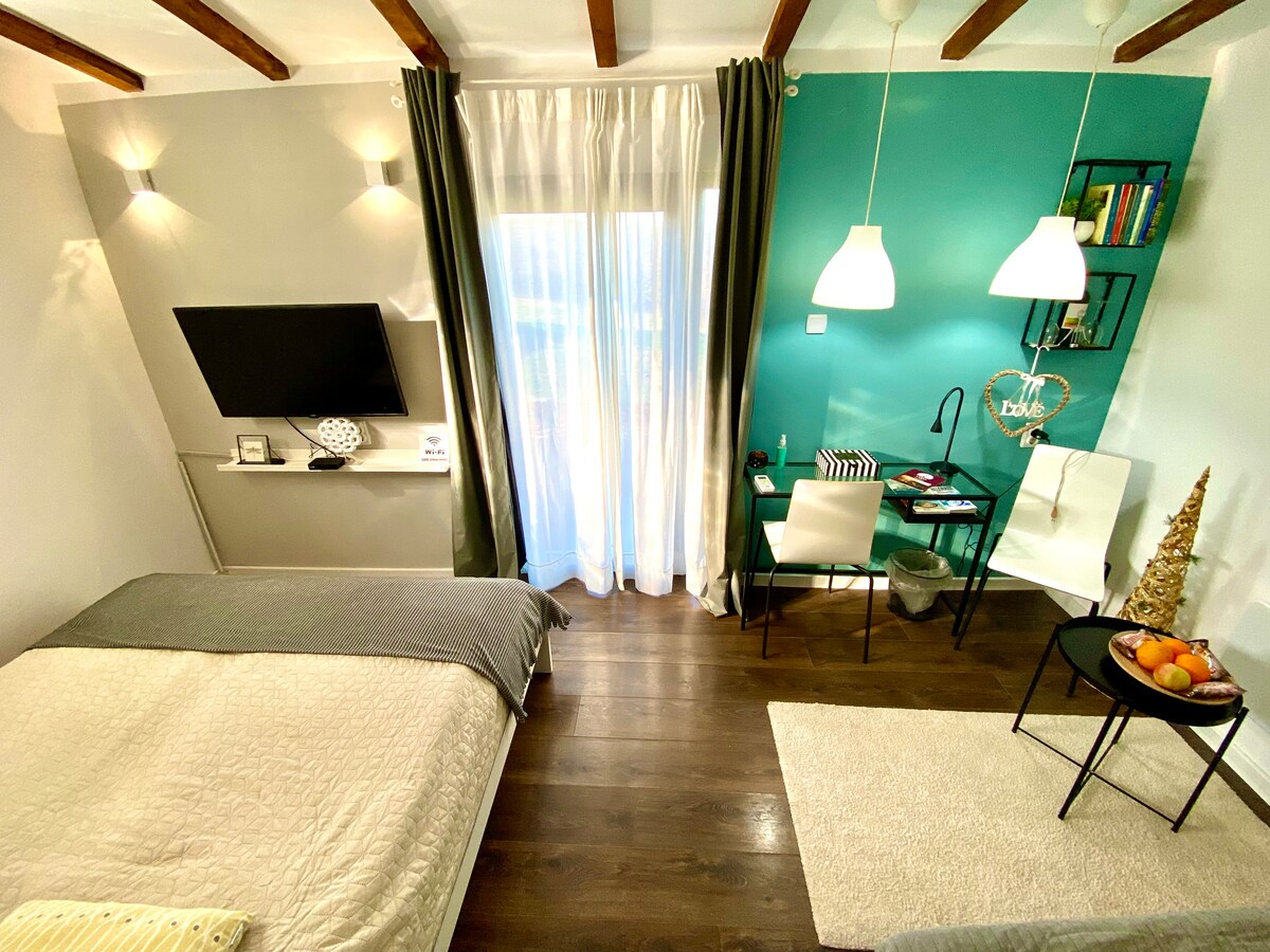 The room features a spacious bed with neutral bedding, complemented by a vibrant teal accent wall. A small desk and chair are positioned near a large window, adorned with sheer curtains. A television is mounted on the wall, and decorative items are placed on a nearby table.