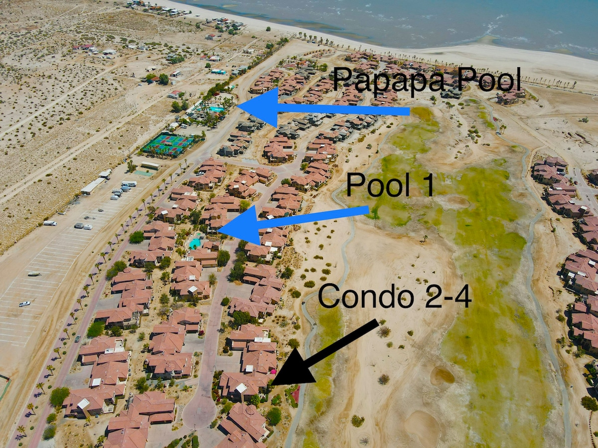 An aerial view captures the layout of the community, highlighting the proximity of various pools and the condo units. The landscape consists of a mix of desert terrain and green golf course areas, with clear markings indicating the locations of the Papapa Pool and Pool 1.