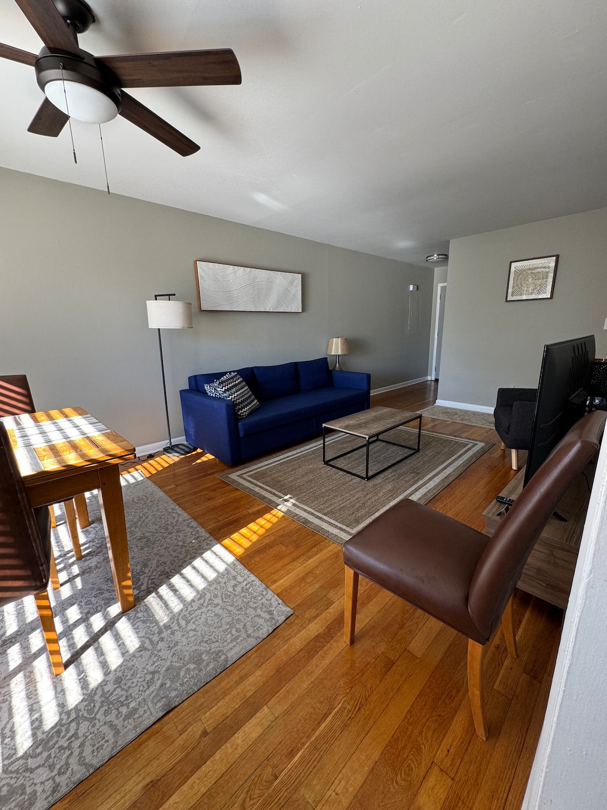 The living area features a blue sofa, two chairs, and a small coffee table arranged on a patterned rug. A dining table with four wooden chairs is illuminated by natural light from the window. A ceiling fan adds a functional touch to the space.