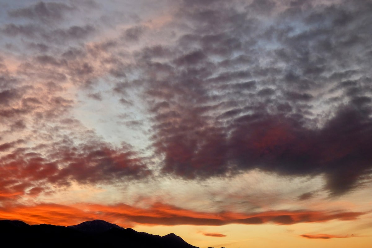 A vibrant sunset sky displays a mix of deep orange and soft purple hues. The silhouettes of mountains create a serene backdrop against the colorful cloud formations, offering a peaceful and picturesque view.