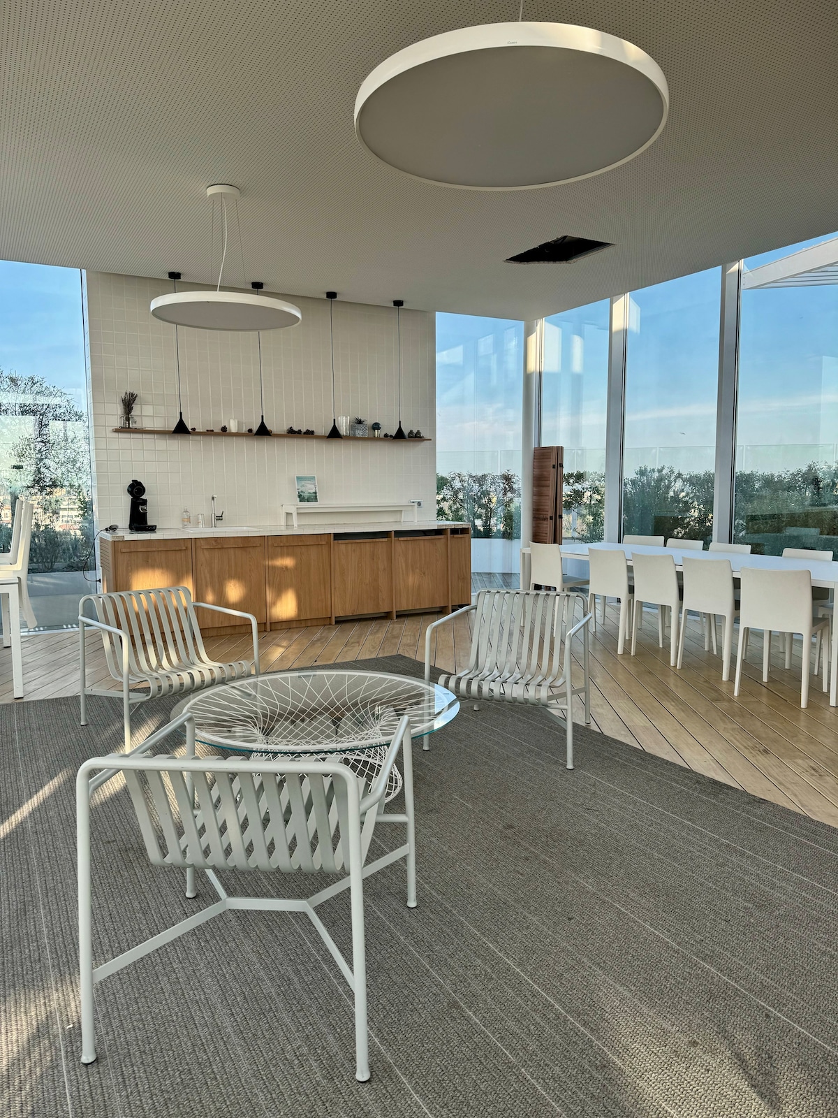 A modern communal space is highlighted by large glass windows that offer abundant natural light. White chairs surround a glass table, complemented by a wooden kitchenette and sleek pendant lighting. The wood flooring contrasts with a neutral area rug, enhancing the area's open and airy feel.