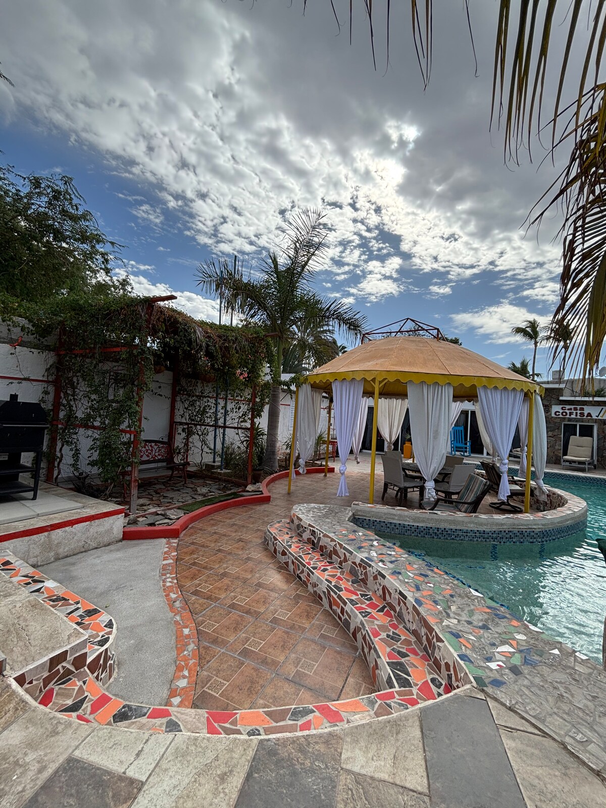 A private pool area is framed by lush greenery and palm trees. A cabana with flowing white curtains provides shade and comfort. The stone pathways lead around the pool, creating an inviting outdoor space for relaxation and enjoyment.