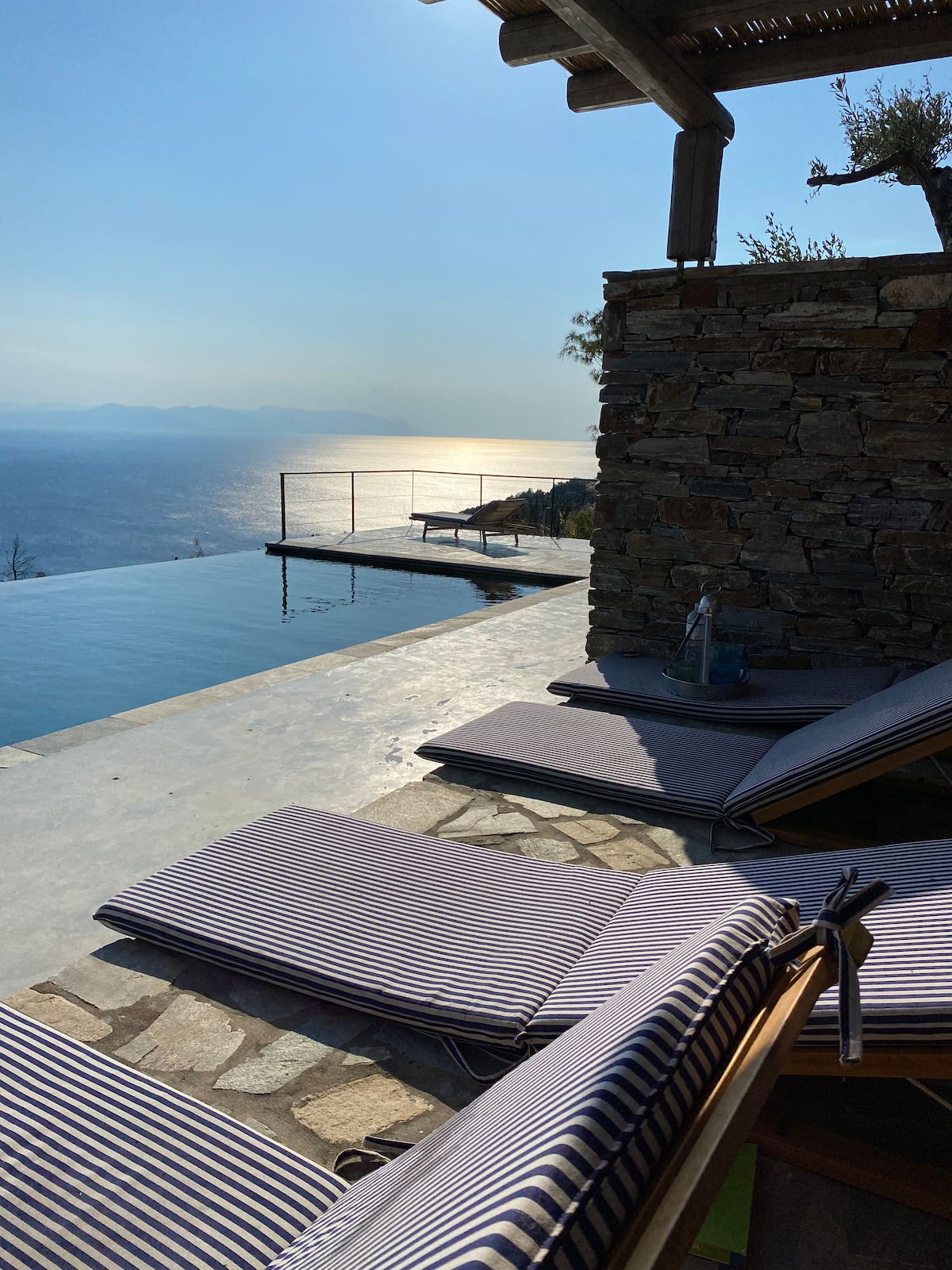A serene infinity pool extends towards the horizon, reflecting the sky and distant land. Lounge chairs with striped cushions are positioned nearby on a stone terrace, offering a relaxing space to enjoy the view. Natural elements blend harmoniously in this tranquil setting.
