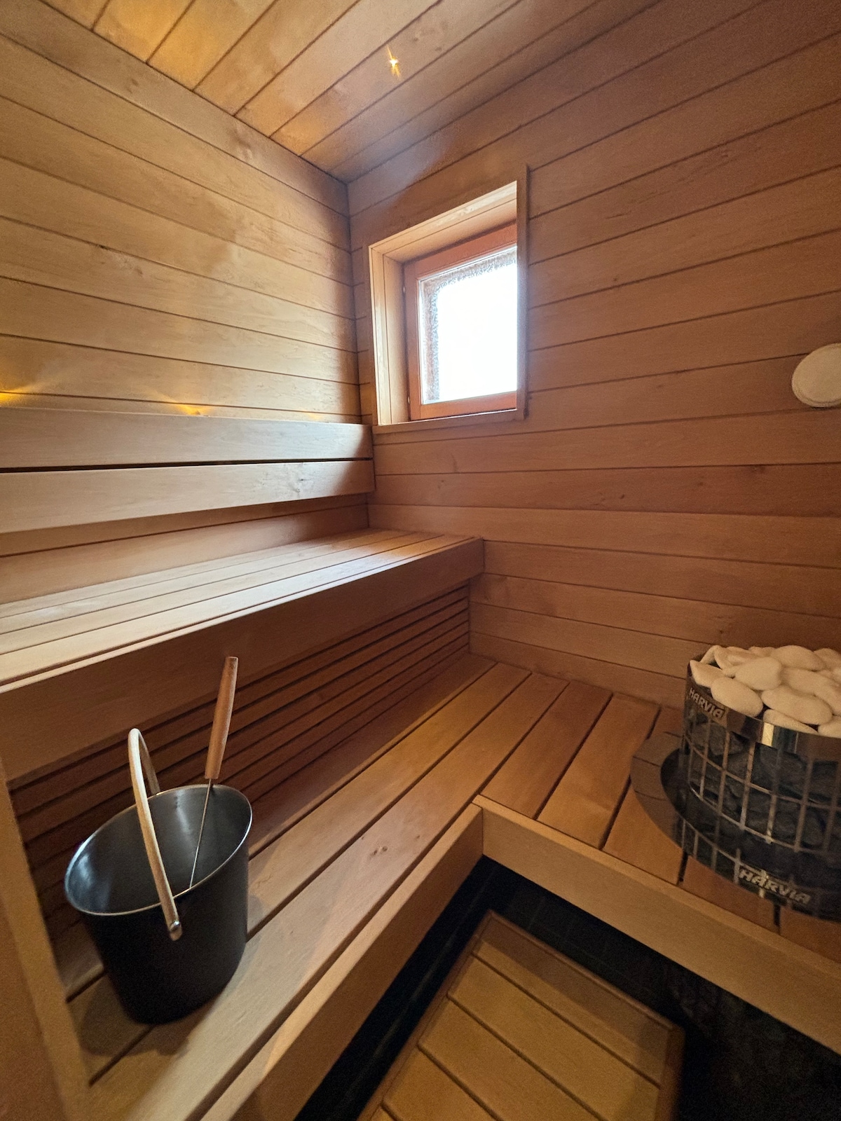 A modern sauna features wooden benches and a window allowing natural light to filter in. A black bucket and a basket filled with towels are visible, enhancing the overall functionality of the space.